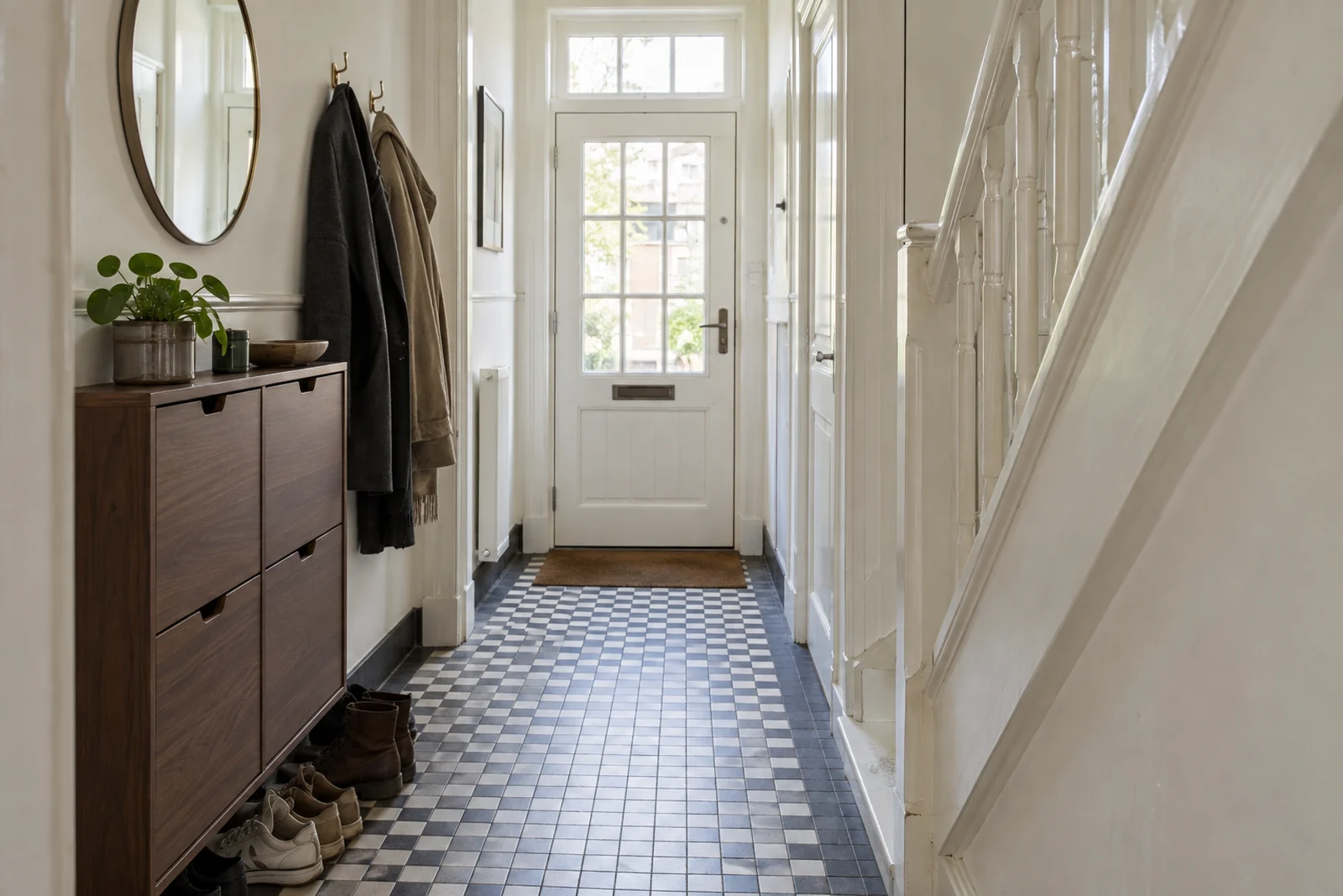 Smalle hal met een witte trap, een houten schoenenkast en een tegelvloer.